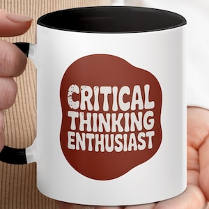 May include: White ceramic mug with a black handle and interior. The mug features the text "CRITICAL THINKING ENTHUSIAST" in white, set against a maroon-coloured, irregular shape. The mug is held by a person.