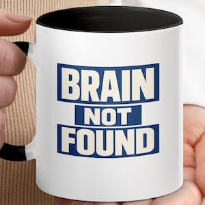 May include: White ceramic mug with a black interior and handle. The mug features the text "BRAIN NOT FOUND" in bold, blue letters on a white background. The mug is held by a person.
