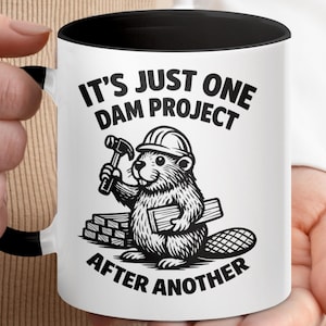 May include: White ceramic mug with a black interior and handle. The mug features a black and white illustration of a beaver wearing a hard hat and holding a hammer and wood planks. Text on the mug reads "IT'S JUST ONE DAM PROJECT AFTER ANOTHER."