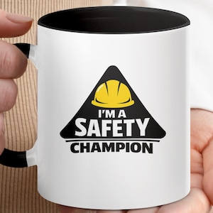 Könnte beinhalten: Ein weißer Keramikbecher mit schwarzem Innenraum und Henkel. Auf der Vorderseite befindet sich ein schwarzes Dreieck mit einem gelben Schutzhelm-Symbol über dem weißen Text "I'M A SAFETY CHAMPION" in einer serifenlosen Schriftart.