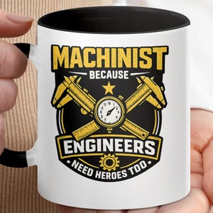 May include: White ceramic mug with a black interior and handle. The mug features a yellow and black design with the words "MACHINIST BECAUSE ENGINEERS NEED HEROES TOO" and a graphic of calipers and a clock.