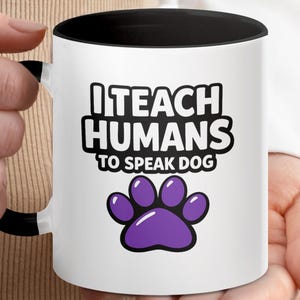Könnte beinhalten: Weißer Keramikbecher mit schwarzem Innenraum und Henkel. Der Becher hat den Text "I TEACH HUMANS TO SPEAK DOG" in Schwarz, mit einem lila Pfotenabdruck darunter. Der Becher wird von einer Person gehalten.