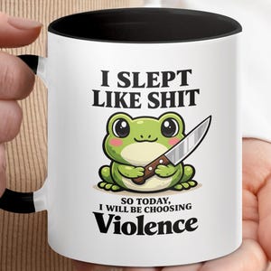 May include: White ceramic mug with a black interior and handle. The mug features the text "I SLEPT LIKE SHIT" above a cartoon frog holding a knife, with the text "SO TODAY, I WILL BE CHOOSING Violence" below.