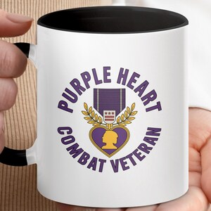 May include: White ceramic mug with a black interior and handle. The mug features a purple and gold design with the words "PURPLE HEART COMBAT VETERAN" encircling a Purple Heart medal graphic. The mug is held by a person.
