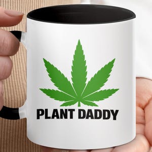 Puede incluir: Taza de cerámica blanca con interior y asa negros. La taza presenta un gran gráfico de hoja de cannabis verde y las palabras "PLANT DADDY" en negro y negrita.