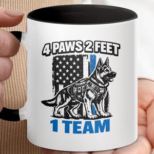 Puede incluir: Una taza de cerámica blanca con asa y borde negros, que presenta un gráfico de un pastor alemán con chaleco táctico frente a una bandera estadounidense desgastada con una franja azul. El texto dice 4 PAWS 2 FEET 1 TEAM.