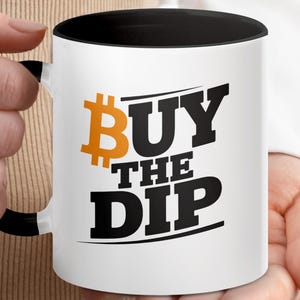 Puede incluir: Taza de cerámica blanca con interior y asa negros. La taza presenta el texto "BUY THE DIP" en letras negras en negrita, con un símbolo de Bitcoin naranja. La taza está sostenida por una persona.