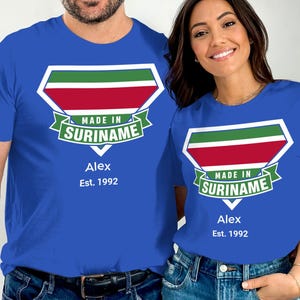 Könnte beinhalten: Zwei Personen tragen passende blaue T-Shirts. Jedes Shirt hat ein schildförmiges Design mit grünen, roten und weißen Streifen und dem Text "MADE IN SURINAME". Darunter stehen der Name "Alex" und "Est. 1992".