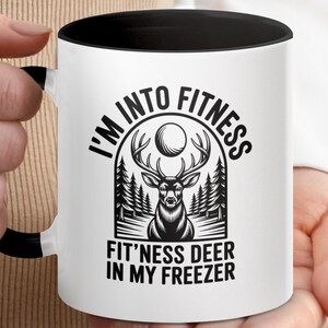 Könnte beinhalten: Weißer Keramikbecher mit schwarzem Innenraum und Griff. Der Becher hat eine schwarz-weiße Grafik mit dem Text "I'M INTO FITNESS" über einer Hirschillustration und dem Text "FIT'NESS DEER IN MY FREEZER".