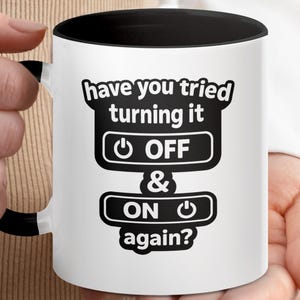 Könnte beinhalten: Ein weißer Keramikbecher mit schwarzem Innenraum und Henkel. Auf der Vorderseite befindet sich der schwarze Schriftzug: have you tried turning it OFF & ON again? Das Design enthält zwei Power-Button-Symbole neben den Wörtern OFF und ON.