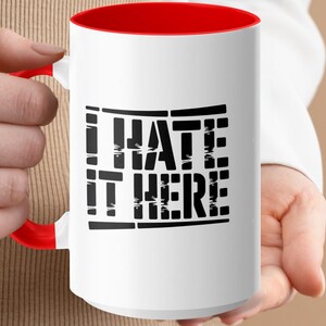 White mug with red handle and "I HATE IT HERE" text.