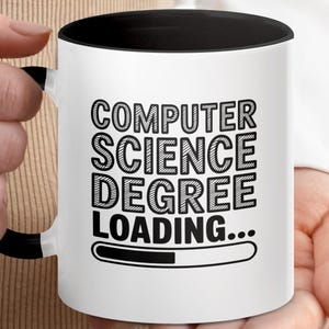 Könnte beinhalten: Weißer Keramikbecher mit schwarzem Innenraum und Henkel. Der Becher zeigt den Text "COMPUTER SCIENCE DEGREE LOADING..." in fetter, schwarzer Schrift. Eine Ladebalken-Grafik befindet sich darunter. Der Becher wird von einer Person gehalten.