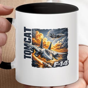 May include: A white ceramic mug with a black interior and handle, featuring a graphic of two F-14 Tomcat fighter jets flying over a sunset ocean scene. The text TOMCAT is printed vertically in dark blue, and F-14 is printed in white at the bottom.