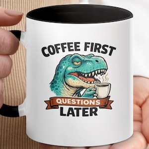 Puede incluir: Taza de cerámica blanca con interior y asa negros. La taza presenta una ilustración de dibujos animados de un dinosaurio turquesa sosteniendo una taza de café, con el texto "COFFEE FIRST QUESTIONS LATER".