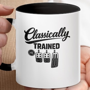 Może przedstawiać: Biały ceramiczny kubek z czarnym wnętrzem i uchwytem. Kubek zawiera napis "Classically Trained" w stylizowanej czcionce, wraz z ilustracjami dźwigni zmiany biegów i pedałów samochodowych. Kubek jest trzymany przez osobę.