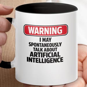 Pode incluir: Uma caneca de cerâmica branca com alça e borda interna pretas. A frente apresenta um sinal de aviso retangular vermelho com a palavra WARNING em branco, seguida pelo texto preto: I MAY SPONTANEOUSLY TALK ABOUT ARTIFICIAL INTELLIGENCE.