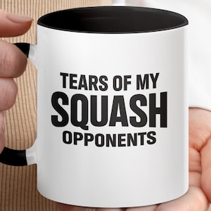 Könnte beinhalten: Weißer Keramikbecher mit schwarzem Innenraum und Griff. Der Becher trägt den Text "TEARS OF MY SQUASH OPPONENTS" in fetten, schwarzen Buchstaben. Der Becher wird von einer Person gehalten.