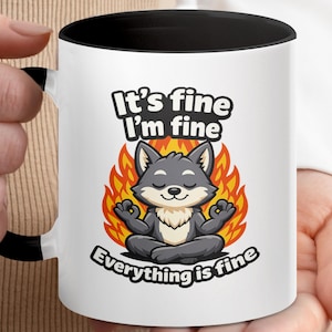 May include: A white ceramic mug with a black interior and handle, featuring a graphic of a grey wolf sitting in a meditative pose in front of orange and yellow flames. The text reads It's fine I'm fine and Everything is fine in black letters.