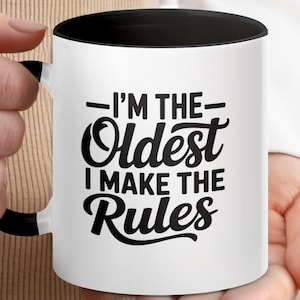 May include: White ceramic mug with a black interior and handle. The mug features the text "I'M THE Oldest I MAKE THE Rules" in a bold, black, stylized font. The mug is held by a person.