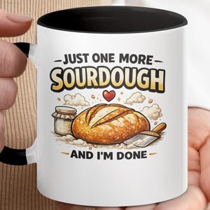 Peut inclure: Mug en céramique blanche avec intérieur et anse noirs, présentant un motif de pain au levain. La tasse porte l'inscription "JUST ONE MORE SOURDOUGH AND I'M DONE" avec des illustrations de pain, d'un pot et d'un cœur.