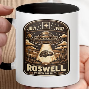May include: White ceramic mug with a black interior and handle, featuring a brown and tan graphic. The graphic depicts a UFO over a car with the text "July 1947 Roswell We Know The Truth."