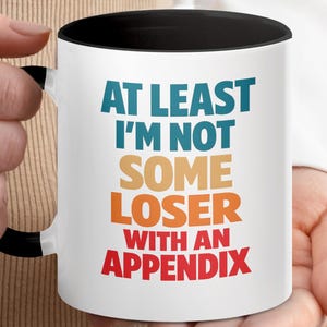 May include: A white ceramic mug with a black handle and interior rim. The front features multi-colored text that reads: AT LEAST I'M NOT SOME LOSER WITH AN APPENDIX. The words are stacked in blue, tan, orange, and red, creating a bold graphic design.