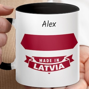 May include: White ceramic mug with a black interior and handle, featuring the name "Alex" in black text. The mug has a burgundy arrow-shaped design and a banner that reads "MADE IN LATVIA" in white text.
