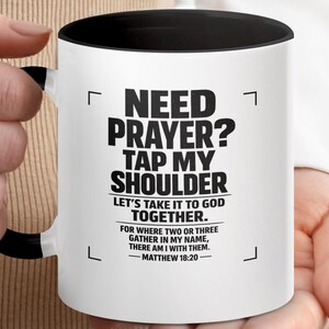 Può includere: Tazza in ceramica bianca con interno e manico neri. La tazza presenta il testo "NEED PRAYER? TAP MY SHOULDER" in grassetto nero, insieme a un versetto biblico. Un regalo ideale per chi cerca conforto o fede.