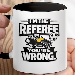 May include: White ceramic mug with a black interior and handle. The mug features the text "I'M THE REFEREE YOU'RE WRONG." along with a whistle and soccer ball graphic. The design is in black and yellow.