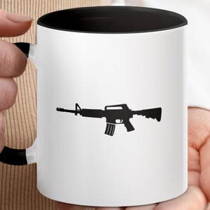 Pode incluir: Caneca de cerâmica branca com interior e alça pretos. A caneca apresenta uma silhueta preta de um rifle. A caneca é segurada por uma pessoa.