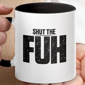 May include: A white ceramic mug with a black interior and a black handle. The front of the mug features the text SHUT THE FUH in bold, distressed black lettering, with the word SHUT THE positioned above the large, prominent letters FUH.