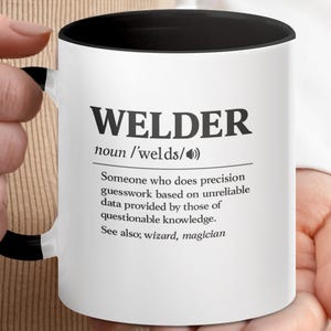 May include: White ceramic mug with a black interior and handle, featuring the word "WELDER" in bold black letters. The mug includes a definition of a welder, with the words "wizard, magician" listed at the end.