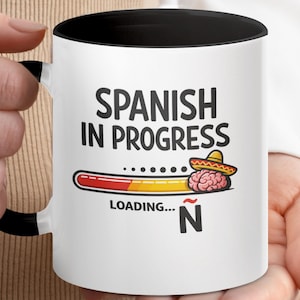 Może przedstawiać: Biały ceramiczny kubek z czarnym wnętrzem i uchwytem. Kubek zawiera napis "SPANISH IN PROGRESS" nad grafiką paska ładowania z mózgiem w sombrero. Wyświetlane jest również słowo "LOADING..." i litera "Ñ".