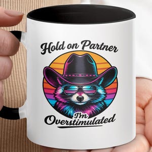 Op de afbeelding: Witte keramische mok met een zwarte handgreep en binnenkant. De mok heeft een kleurrijke illustratie van een wasbeer met een cowboyhoed en zonnebril, met de tekst "Hold on Partner, I'm Overstimulated."