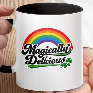 Könnte beinhalten: Weißer Keramikbecher mit schwarzem Innenraum und Griff. Der Becher zeigt einen Regenbogen über den Worten "Magically Delicious" in einer skurrilen Schriftart, mit einem grünen Kleeblatt auf der rechten Seite. Ein lustiges und farbenfrohes Design.