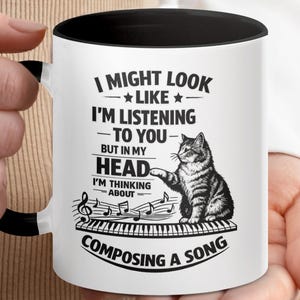 Può includere: Tazza in ceramica bianca con interno e manico neri. La tazza presenta un'illustrazione in bianco e nero di un gatto seduto su una tastiera di pianoforte, con note musicali e il testo "I might look like I'm listening to you but in my head I'm thinking about composing a song."