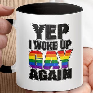 Pode incluir: Uma caneca de cerâmica branca com alça e borda interna pretas. A frente apresenta o texto preto YEP I WOKE UP e AGAIN, com a palavra GAY em letras grandes e em negrito nas cores do arco-íris. O design tem uma aparência texturizada e desgastada.