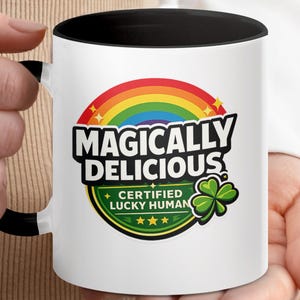 Könnte beinhalten: Weiße Keramik-Tasse mit schwarzem Innenraum und Griff, mit dem Text "MAGICALY DELICIOUS" über einem Regenbogen und einem grünen Kleeblatt. Die Tasse enthält auch die Worte "CERTIFIED LUCKY HUMAN".