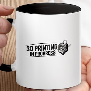 May include: A white ceramic mug with a black interior and a black handle. The front of the mug features a black graphic design with the text 3D PRINTING IN PROGRESS and an illustration of a 3D printer nozzle assembly with a cooling fan.