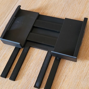 May include: Black plastic phone stand with three adjustable legs. The stand has a slot for a phone and a sliding mechanism to adjust the height.