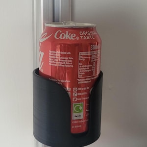 以下が含まれることがあります： 白い壁に取り付けられた黒いプラスチック製の缶ホルダーに、赤いコカ・コーラの缶が入っています。缶は330mlで、「オリジナルテイスト」の文字が書かれています。