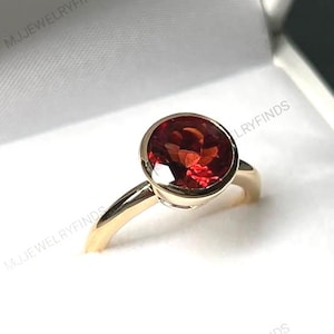Op de afbeelding: Een gouden ring met een grote, ronde, rode edelsteen in een bezel setting. De ring wordt gepresenteerd in een witte sieradendoos. De edelsteen heeft een facet geslepen uiterlijk. Een klassiek sieraad.