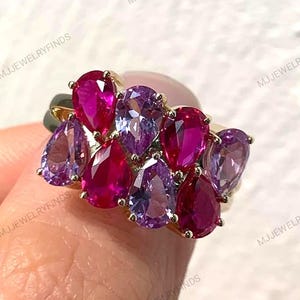 Könnte beinhalten: Ein goldener Ring mit einer Ansammlung von birnenförmigen Edelsteinen in Rosa- und Lilatönen. Das Design des Rings zeigt eine lebendige Anordnung von Steinen, die einen auffälligen visuellen Reiz erzeugt. Der Ring wird von einer Person gehalten.