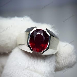 Pu&ograve; includere: Un anello d'argento con una grande pietra preziosa rosso scuro a taglio ovale. La pietra &egrave; incastonata in un design geometrico moderno con accenti neri. L'anello &egrave; mostrato su sfondo bianco.