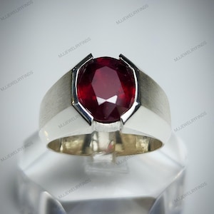 Pu&ograve; includere: Un anello d'argento con una grande pietra preziosa rosso scuro a taglio ovale. L'anello ha una finitura in metallo spazzolato sulla fascia e un'incastonatura in metallo bianco lucido attorno alla pietra preziosa. L'anello &egrave; esposto su una superficie trasparente e angolata.