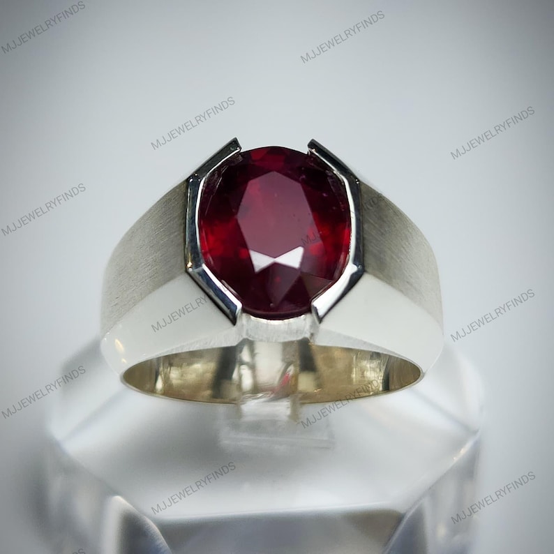 Pu&ograve; includere: Un anello d'argento con una grande pietra preziosa rosso scuro a taglio ovale. L'anello ha una finitura in metallo spazzolato sulla fascia e un'incastonatura in metallo bianco lucido attorno alla pietra preziosa. L'anello &egrave; esposto su una superficie trasparente e angolata.