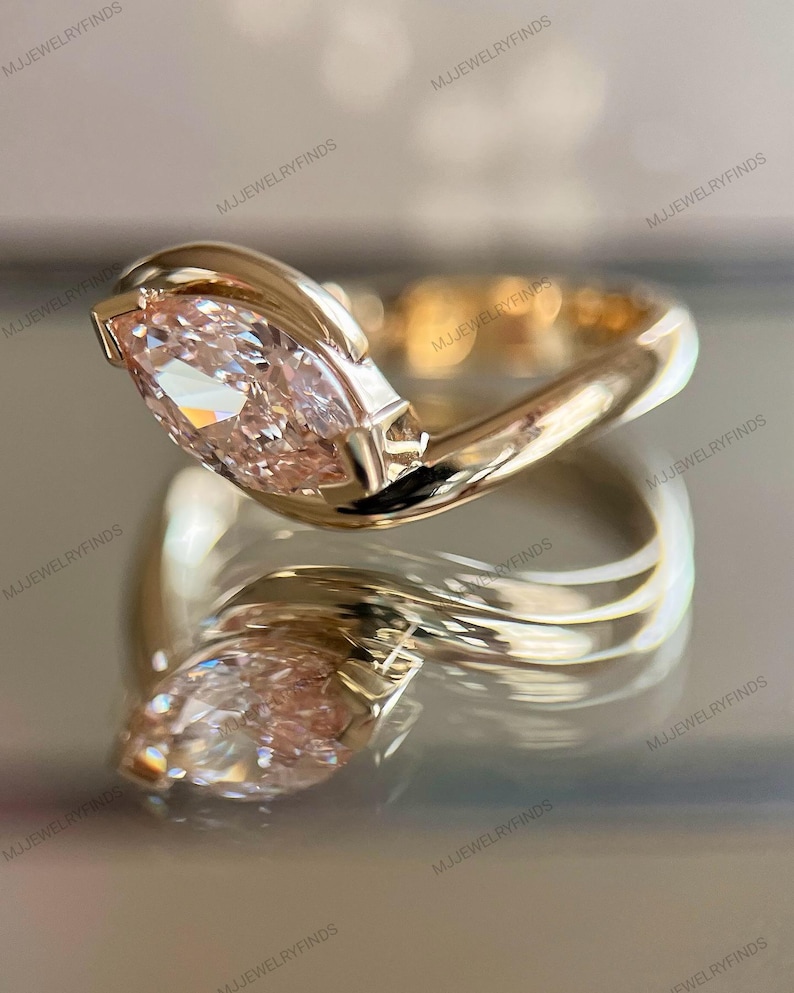 May include: A gold ring with a marquise-cut, light pink gemstone. The ring's band has a unique, flowing design. The ring is highly reflective, with the text "MJJEWELRYFINDS" visible in the background.