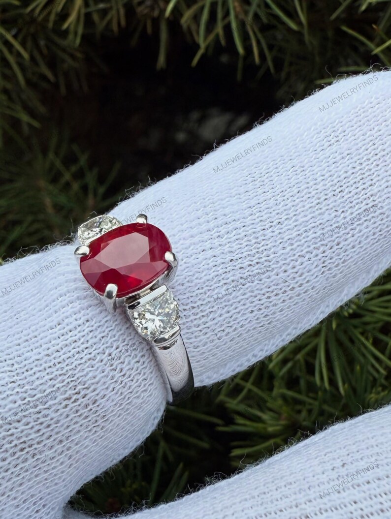 May include: A silver-toned ring featuring a large, oval-cut, deep red ruby gemstone at the center, flanked by two smaller, round-cut, clear white diamonds on either side, all set in a polished metal band against a white fabric background.
