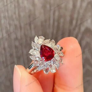 Puede incluir: Un anillo de plata con una gran gema roja oscura en forma de pera en el centro, rodeada por un halo de gemas claras más pequeñas. El diseño del anillo se asemeja a una flor, con la piedra roja como punto focal.