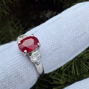 May include: A silver-toned ring featuring a large, oval-cut, deep red ruby gemstone at the center, flanked by two smaller, round-cut, clear white diamonds on either side, all set in a polished metal band against a white fabric background.